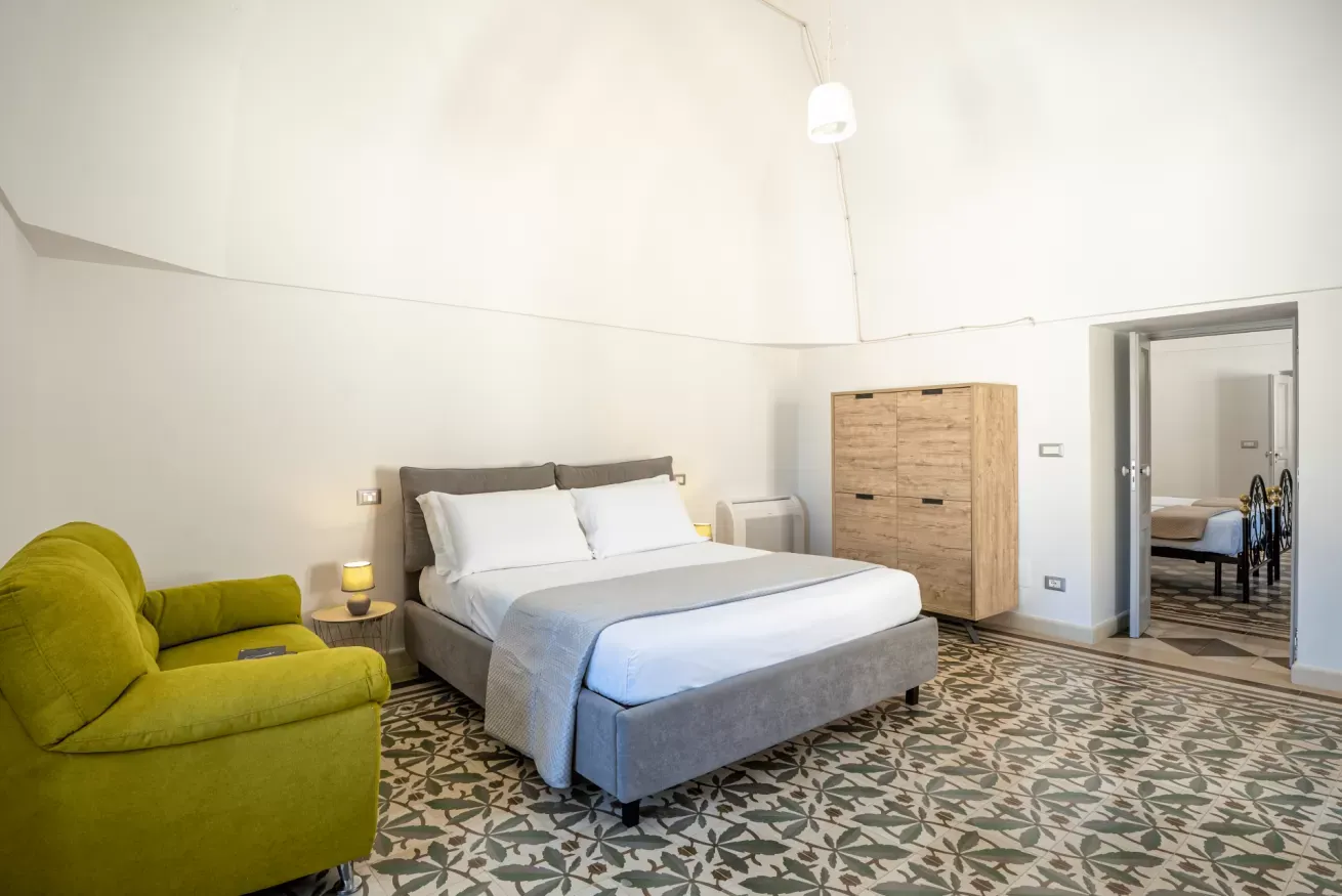 Camera da letto con letto matrimoniale, poltrona verde, comodino, armadio e porta aperta verso un’altra stanza