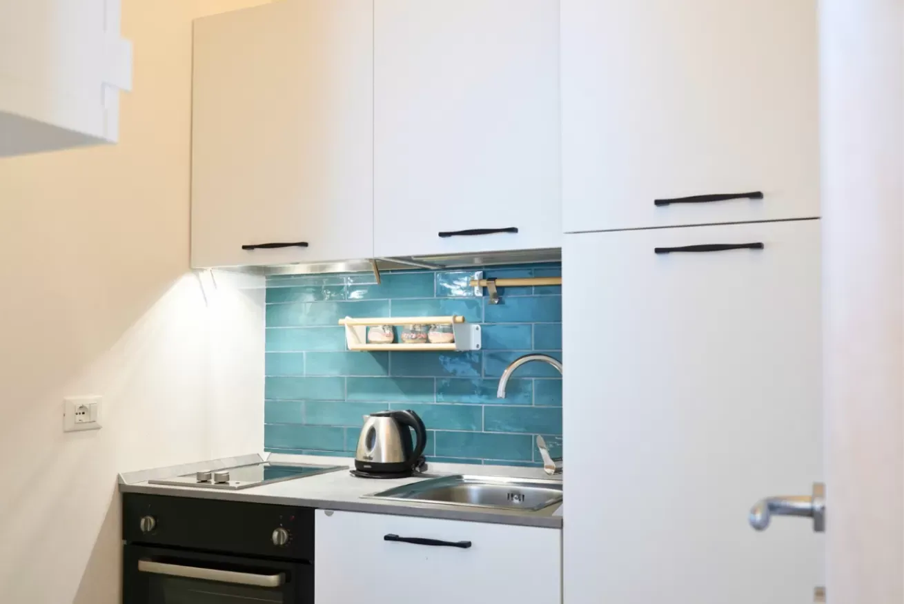 Cucina compatta con pensili bianchi, piastrelle turchesi, piano cottura, lavello e bollitore elettrico