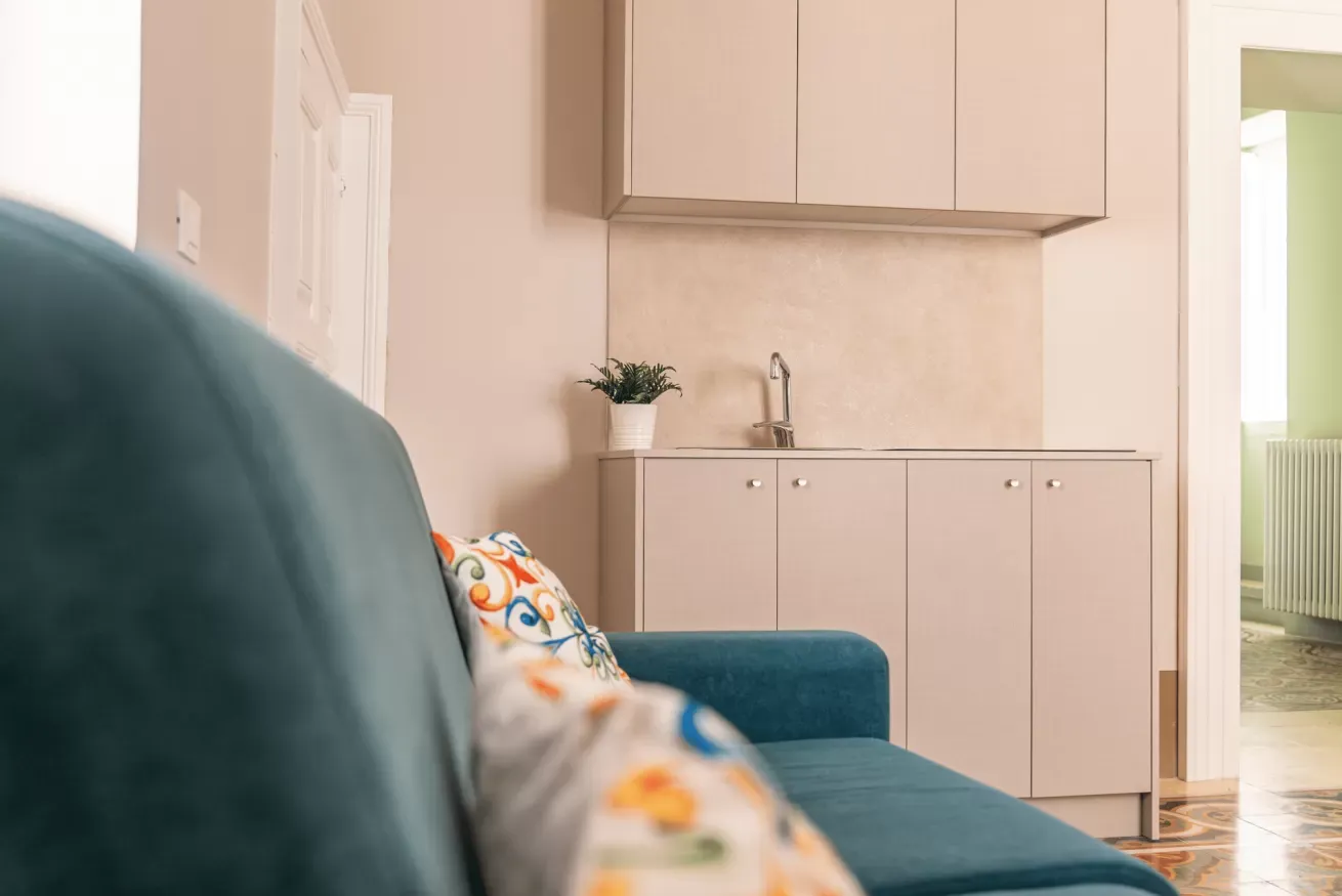 Salotto con divano blu, cuscini decorati e angolo cucina con mobile e lavello