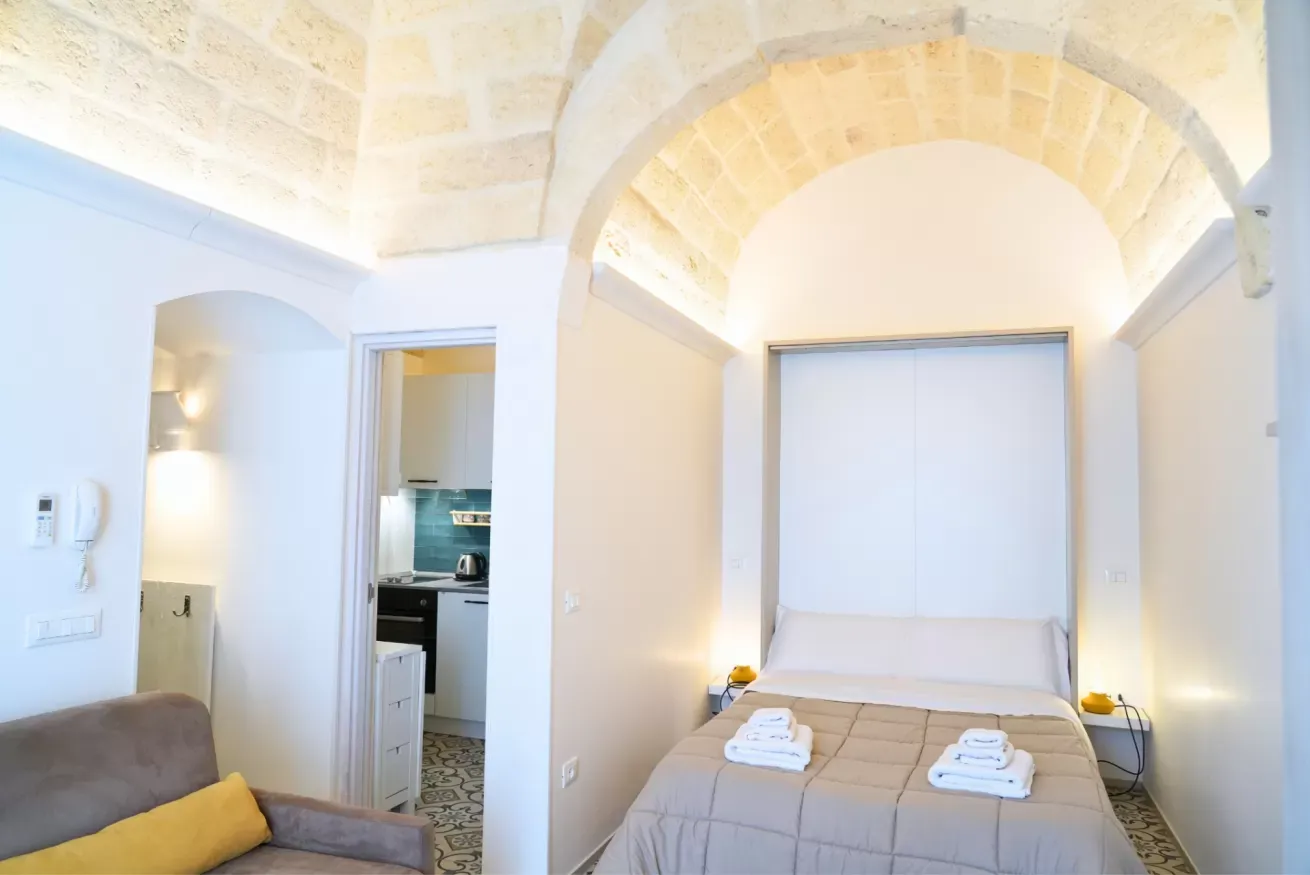 Camera da letto luminosa con letto matrimoniale, arco in pietra e angolo cucina sullo sfondo