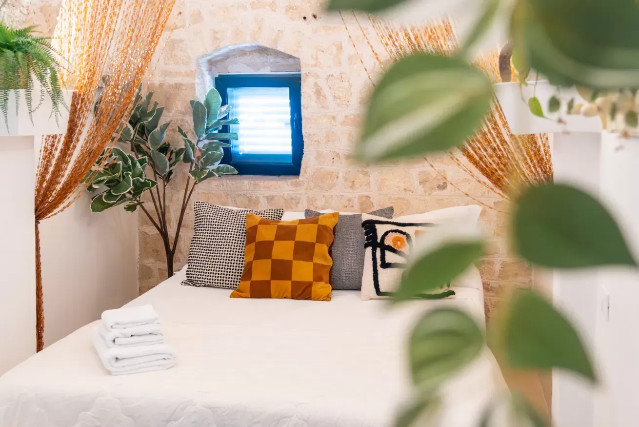 Camera da letto accogliente con letto bianco, cuscini decorativi, piante e finestra azzurra su parete in pietra
