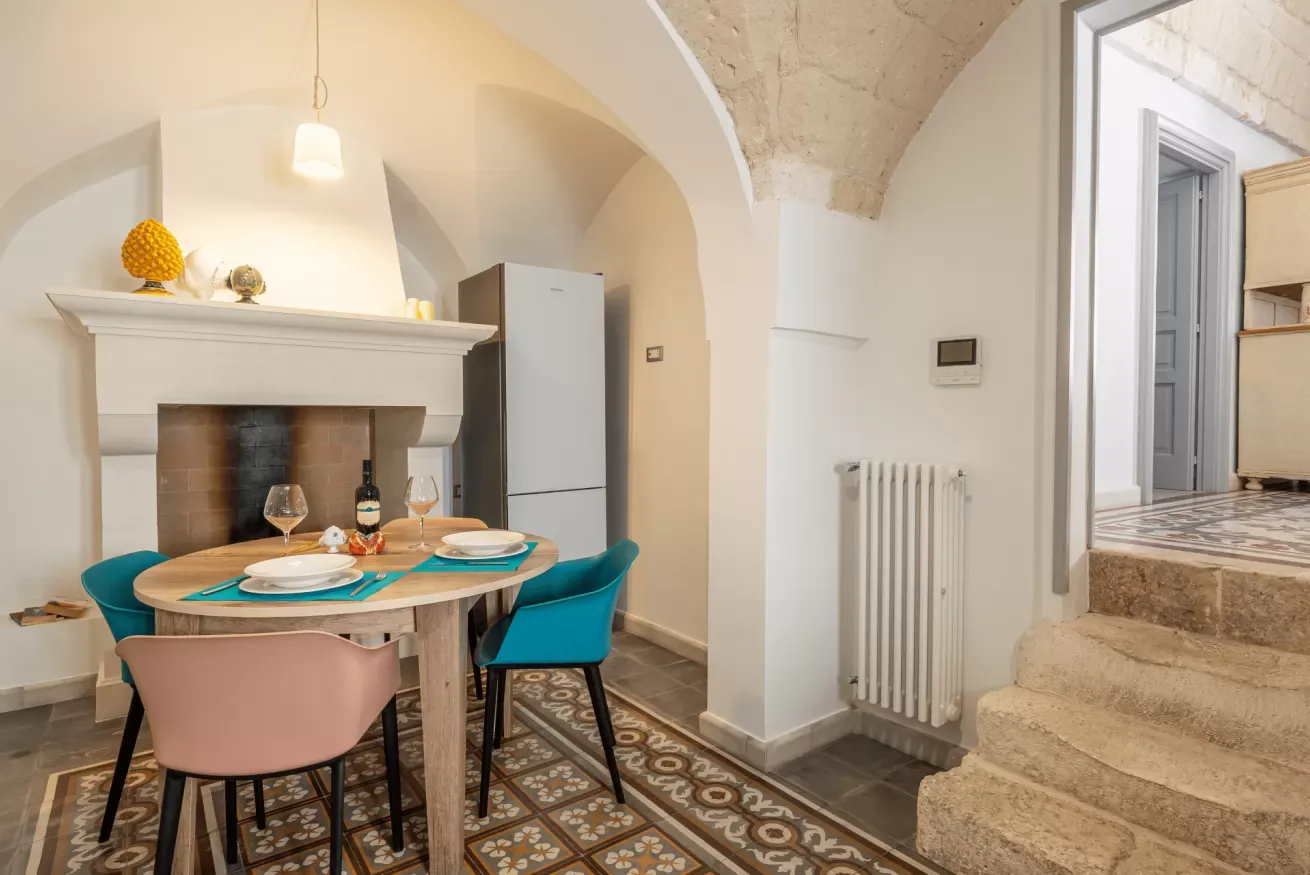 Sala da pranzo con tavolo apparecchiato, camino, frigorifero e scala in pietra sotto volte in muratura